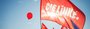 Fahne der Partei DIE LINKE vor einem roten Luftbalon, der vor blauem Himmel aufsteigt.