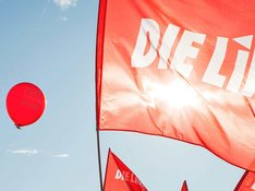 Fahne der Partei DIE LINKE vor einem roten Luftbalon, der vor blauem Himmel aufsteigt.