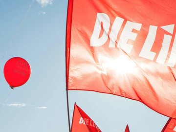 Fahne der Partei DIE LINKE vor einem roten Luftbalon, der vor blauem Himmel aufsteigt.